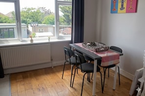A dining area with a table, chairs, a radiator, and a window with a view of buildings and trees. - Spacious 3BR Flat Sleeps 7 in Greenford (Greenford)