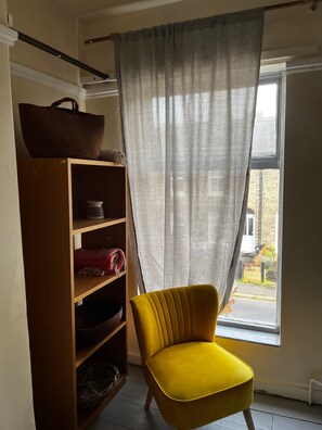 Ein Zimmer mit einem gelben Sessel, einem Holzregal und einem Fenster mit Blick auf ein Backsteinhaus.