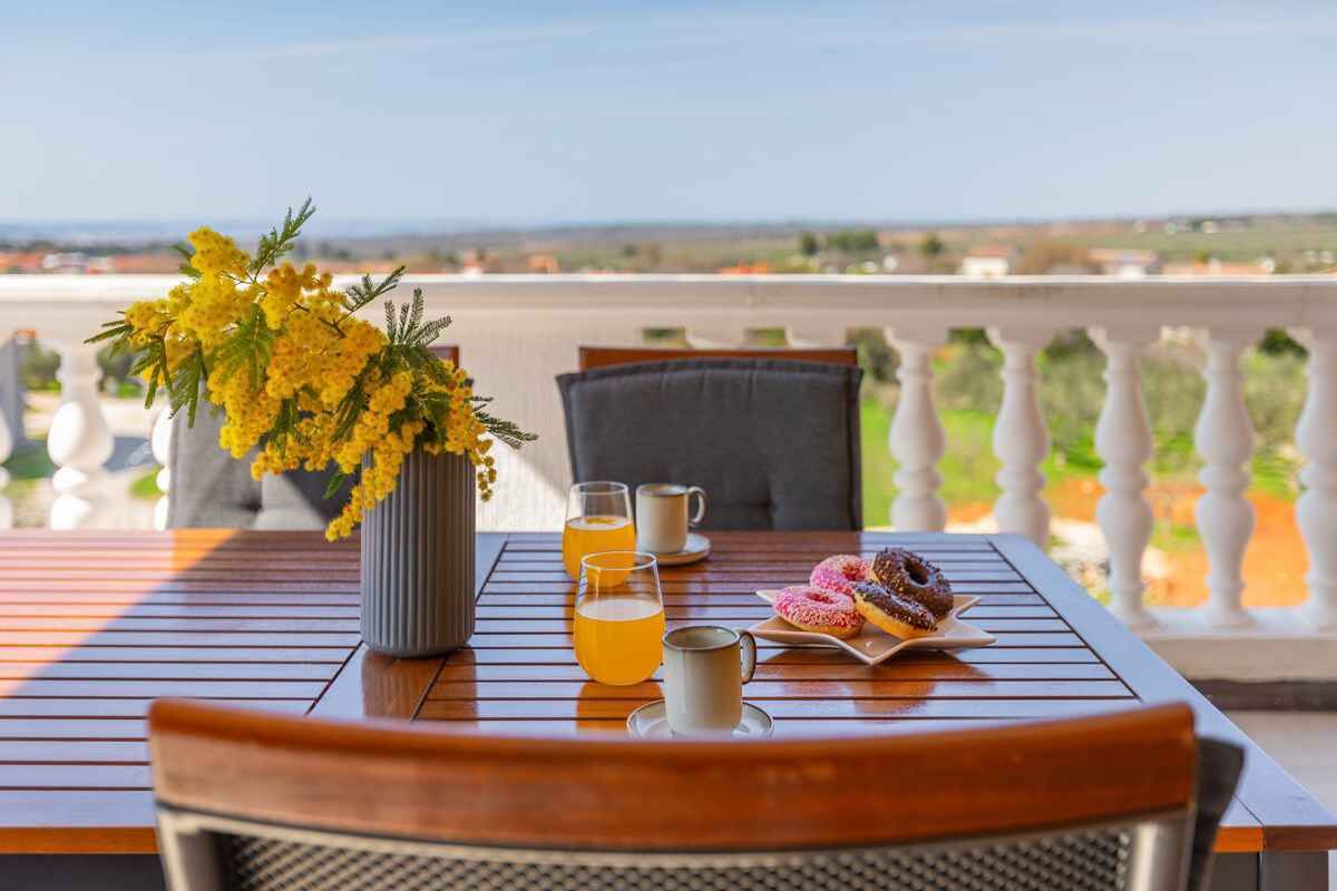Ein Tisch mit einer Vase voller gelber Blumen, zwei Gläser mit orangefarbener Flüssigkeit, einer Tasse und einem Teller mit Donuts auf einem Balkon mit Blick auf eine Landschaft.