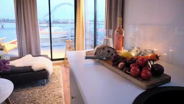 Ett modernt hotellrum med ett stort fönster med utsikt över en sjö eller ett annat vattendrag och en bro. Det finns en matplats med ett bord dukat för två personer, en soffa och ett litet runt bord.