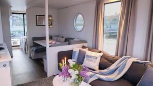 Ett kompakt vardagsrum med en soffa, ett litet bord med en vas med blommor, en rund spegel och utsikt över omgivningarna.