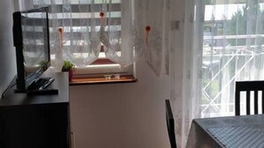 Ein Zimmer mit Esstisch, Stühlen, einem Fernseher auf einem Schrank, einem Fenster mit Jalousien und Blick ins Freie.