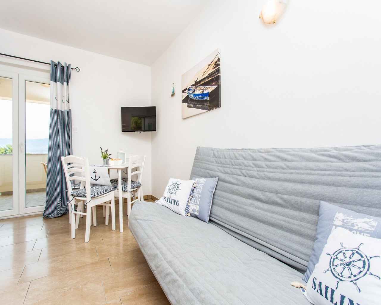 Een woonkamer met een grijze bank, een eettafel met stoelen, een aan de muur bevestigde televisie, en een balkon met gordijnen.