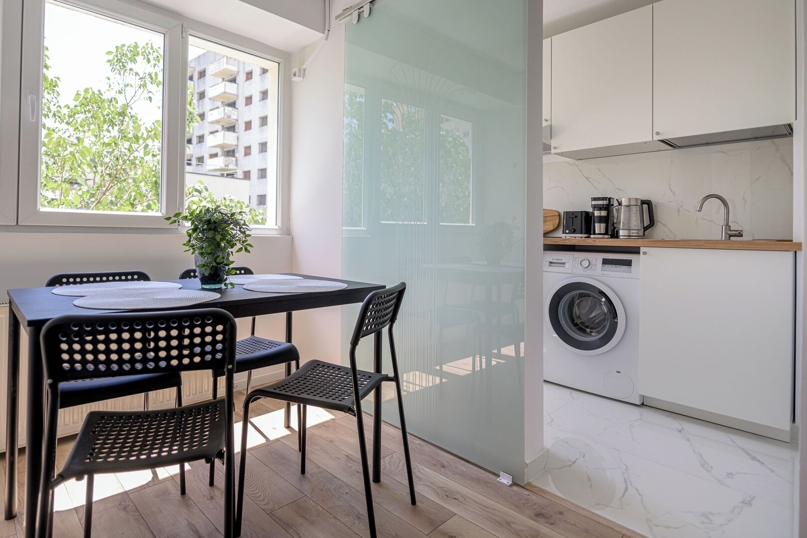 A compact kitchen with a washing machine, a dining area with a table and chairs, and a view of a building outside.