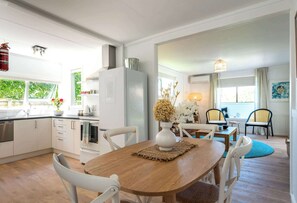 A modern kitchen with white cabinets, a wooden dining table, and a view of the outdoors. - Bach 14 – Relaxed Family Escape in Blackpool (Waiheke Island)