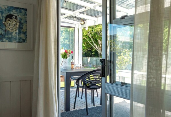 A conservatory with a glass door leading to an outdoor deck, a table with chairs, and a framed artwork on the wall. - Bach 14 – Relaxed Family Escape in Blackpool (Waiheke Island)