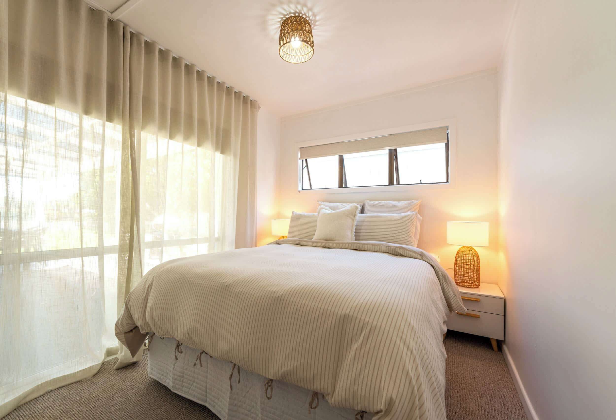 Un dormitorio con cama, mesita de noche con lámpara, ventana con cortinas e iluminación en el techo.