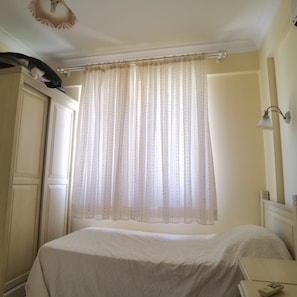 A bedroom with a bed, a nightstand, a lamp, a wardrobe, and a window with a curtain.