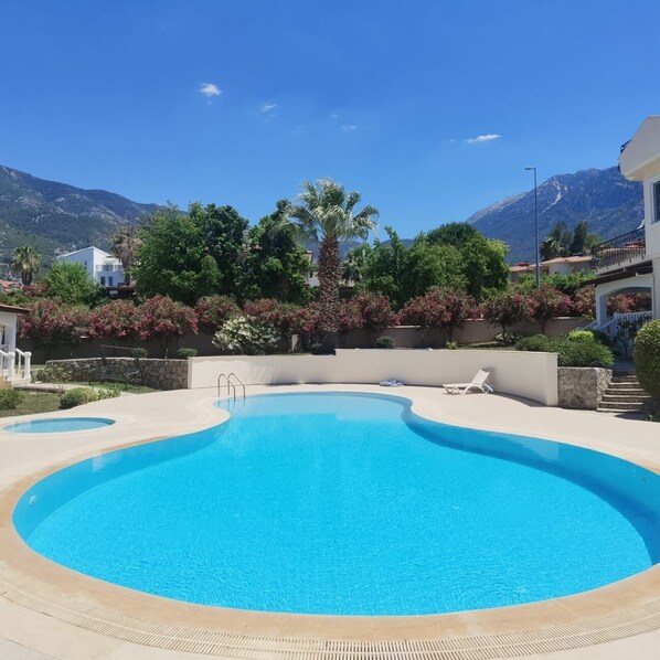 Une piscine à bordures courbes, entourée d’une terrasse en pierre, offrant une vue sur les montagnes et un bâtiment blanc en arrière-plan.