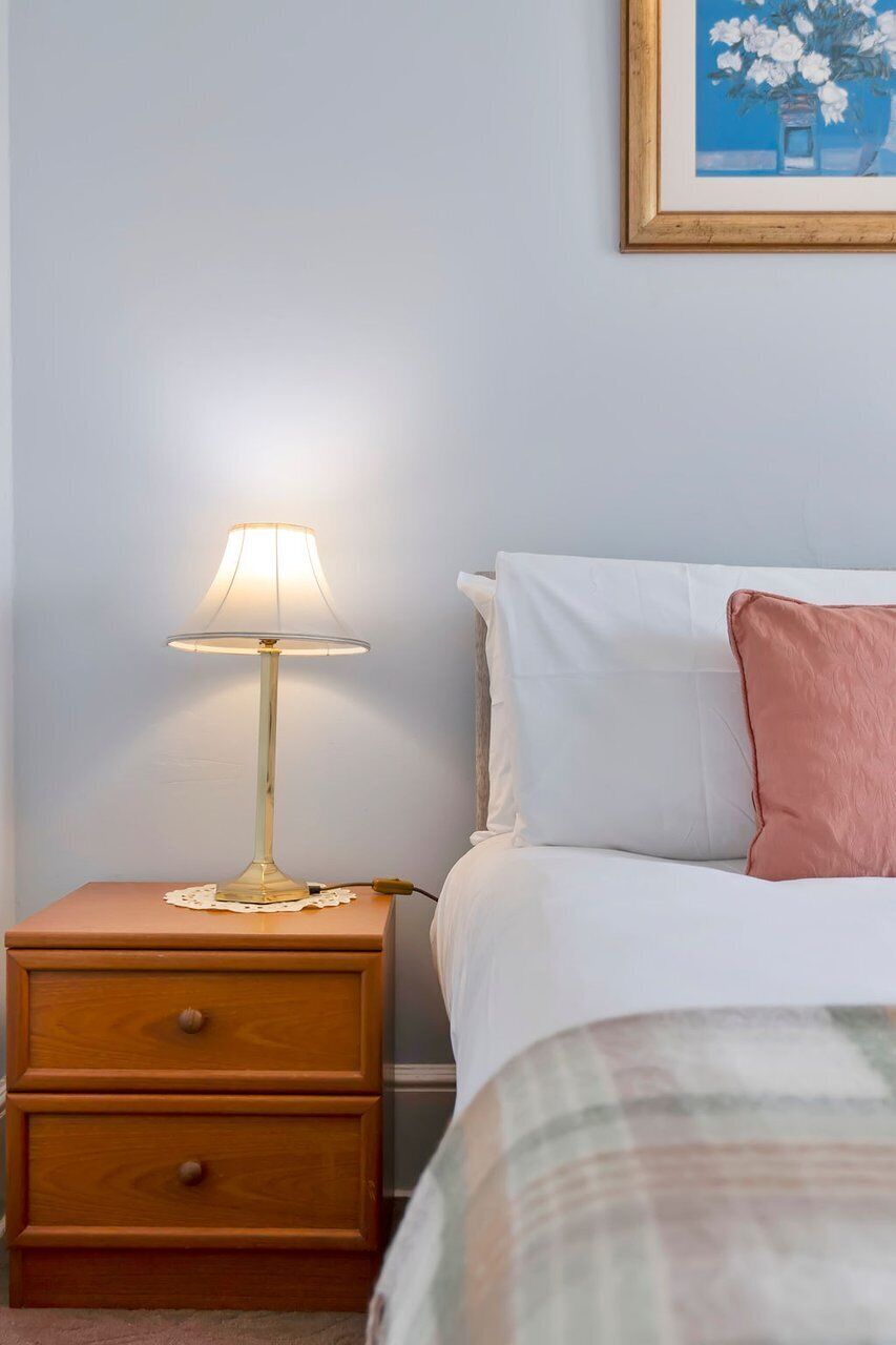 A bedroom with a bedside table, a lamp, a bed with white and pink pillows, and a framed picture on the wall.