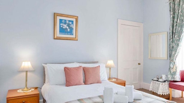 Una camera da letto con un letto, due comodini, un tavolino, un quadro incorniciato, una porta e una finestra con tende.