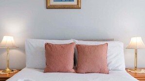 Un letto rifatto con lenzuola bianche e due cuscini rosa, con due lampade ai comodini di legno.