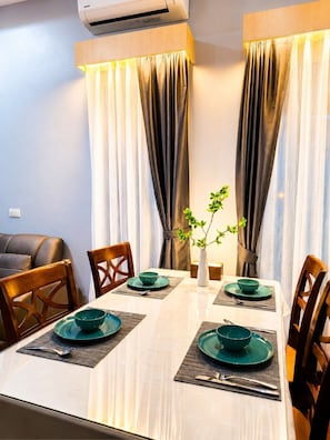 Une salle à manger avec une table dressée pour quatre personnes, deux rideaux, un vase avec une plante et un canapé en cuir.
