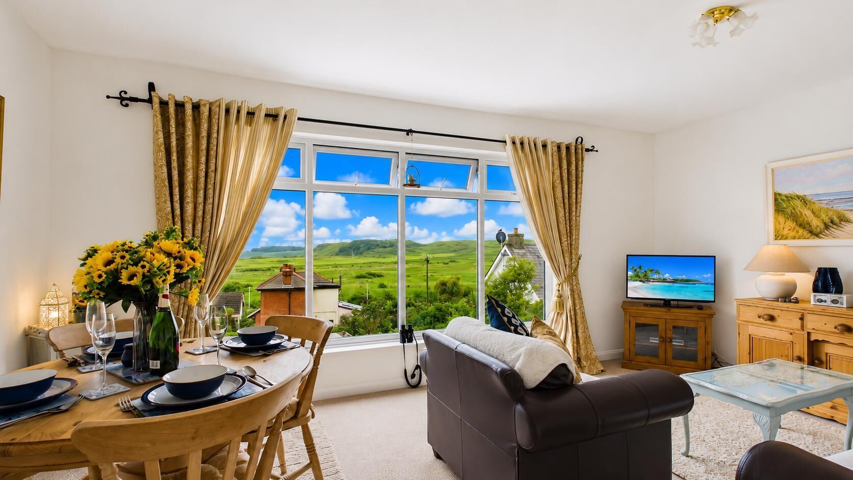 Ein Zimmer mit Esstisch für zwei Personen, einer Couch, einem Fernseher und Blick auf eine grüne Landschaft durch große Fenster.