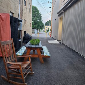 Une ruelle étroite avec une chaise berçante en bois et une table de pique-nique agrémentée d’une plante verte.
