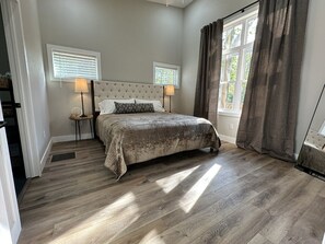 A bedroom with a large bed, two bedside tables, a window with blinds, and a view of greenery outside. - NEW Creekside Home with Dock, Boat Ramp, and Hot Tub (Gallipolis)