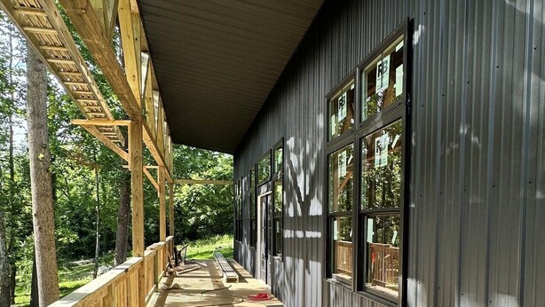 A covered wooden deck with a bench, large windows, and a view of the surrounding forest. - Brand new creekside home with hot tub, private dock and boat ramp! (Gallipolis)
