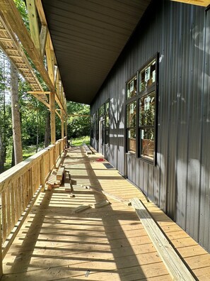 A covered wooden deck with a bench, large windows, and a view of the surrounding forest. - NEW Creekside Home with Dock, Boat Ramp, and Hot Tub (Gallipolis)