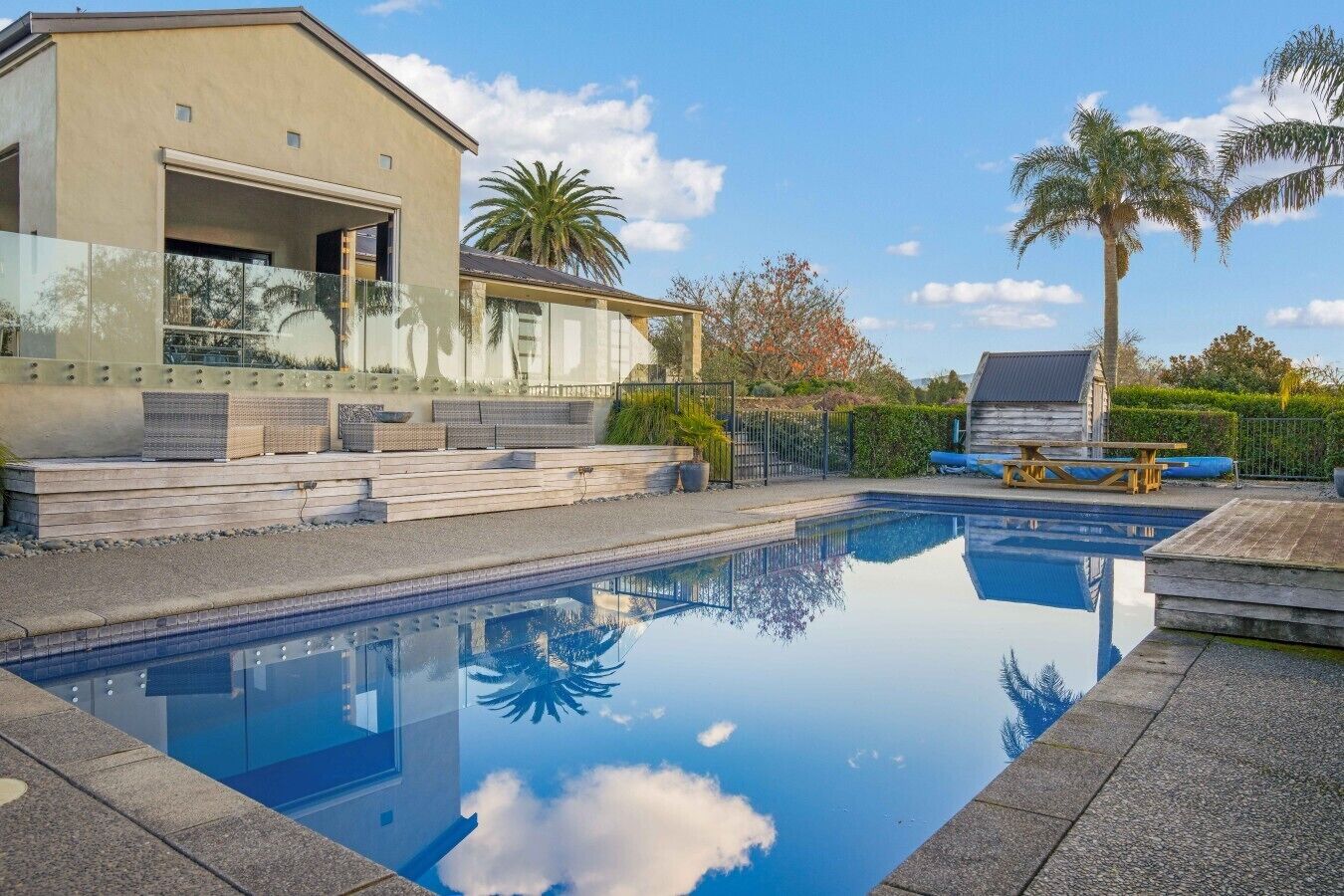 A swimming pool with a clear reflection, a modern house with a garage, and a palm tree in the background.
