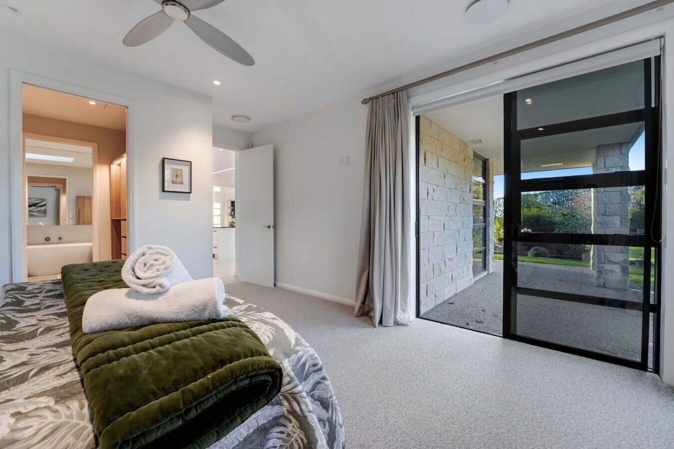 A bedroom with a bed, a ceiling fan, a bathroom visible through an open door, and a sliding glass door leading to an outdoor area.