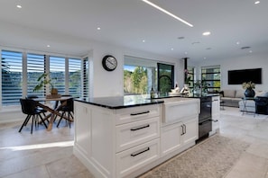 A modern kitchen with white cabinetry, a black countertop, and a dining area with a table and chairs. - Te Puna 5BR Estate | Pool, Tennis, Sauna, Cottage (Tauranga)