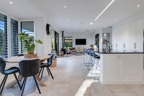 A modern kitchen with a central island, bar seating, and a dining area with chairs and a table. - Te Puna 5BR Estate | Pool, Tennis, Sauna, Cottage (Tauranga)