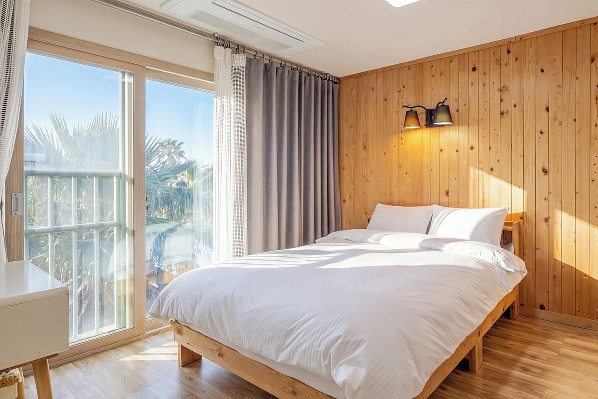 A bedroom with a wooden bed, a desk, a balcony with curtains, and a wall-mounted lamp.