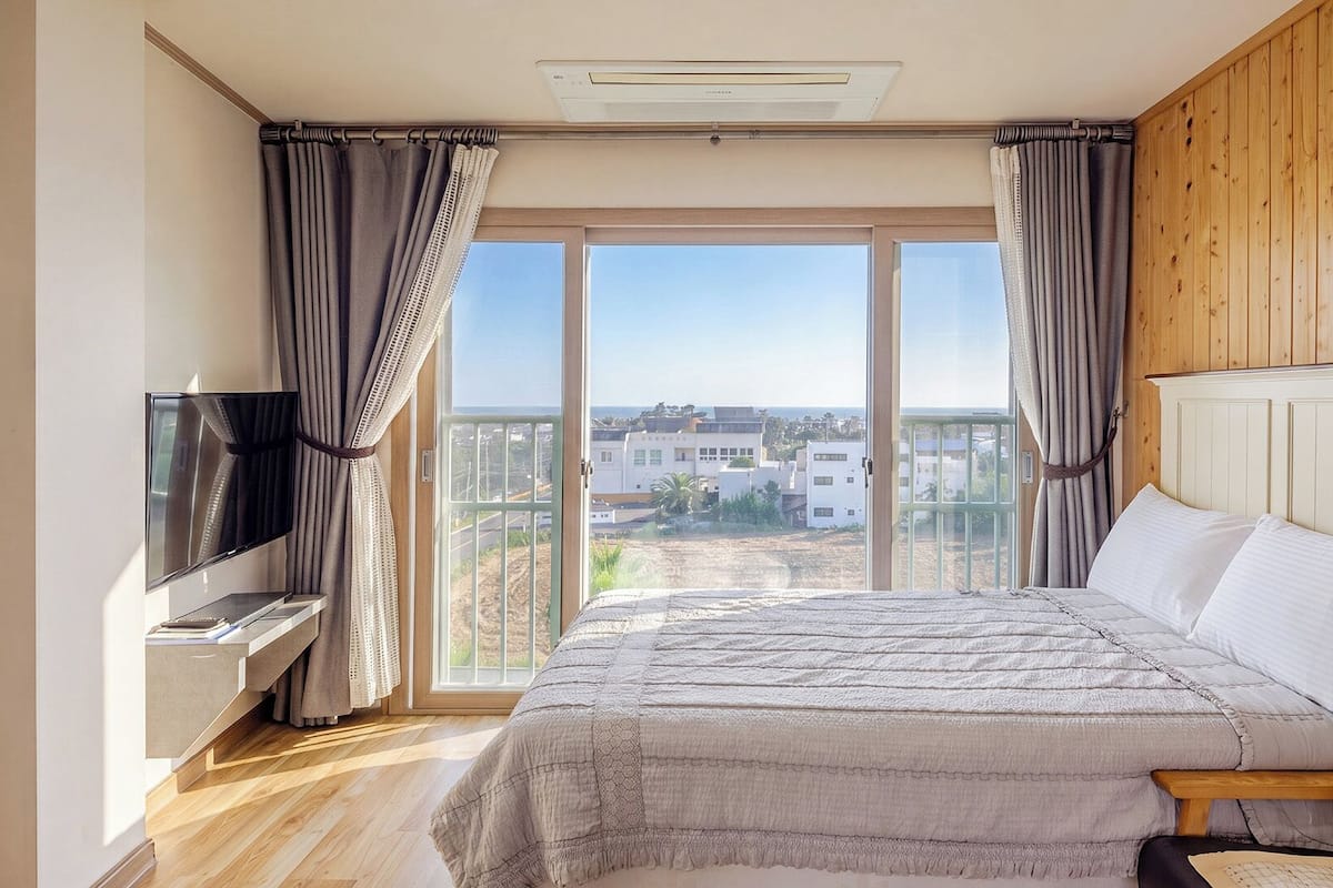 A bedroom with a bed, TV, and a view of buildings through large windows.