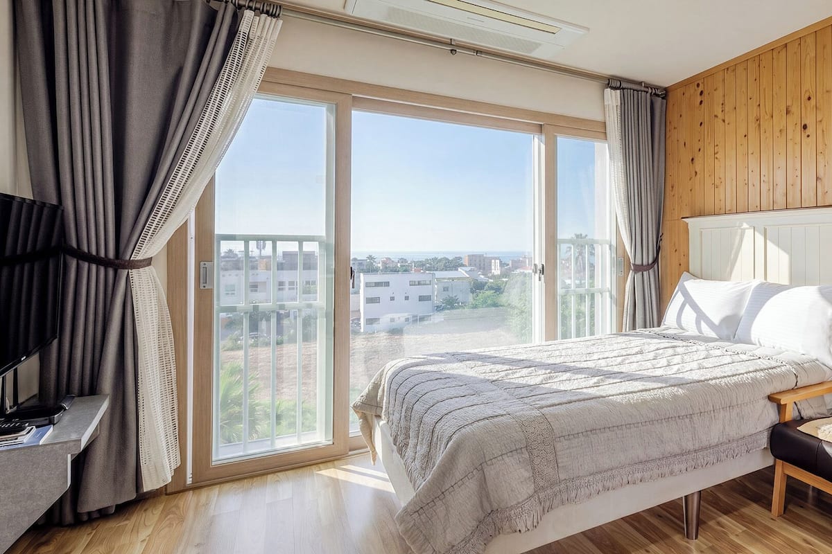 A bedroom with a bed, a sofa, a TV, and a large window with curtains.