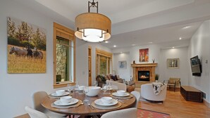 Un comedor con una mesa redonda dispuesta para cuatro personas, rodeada de sillas. Hay una chimenea con repisa y un televisor montado en la pared.
