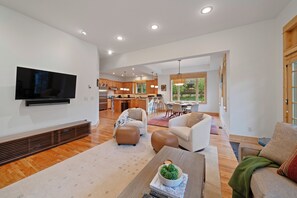 A spacious living area with a flat-screen TV, comfortable seating, a kitchen in the background, and large windows.