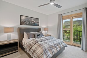 A bedroom with a bed, a headboard, a nightstand, a lamp, a ceiling fan, a window with curtains, and a balcony with a chair. - Town Center Home with Private Hot Tub (Gallatin Gateway)