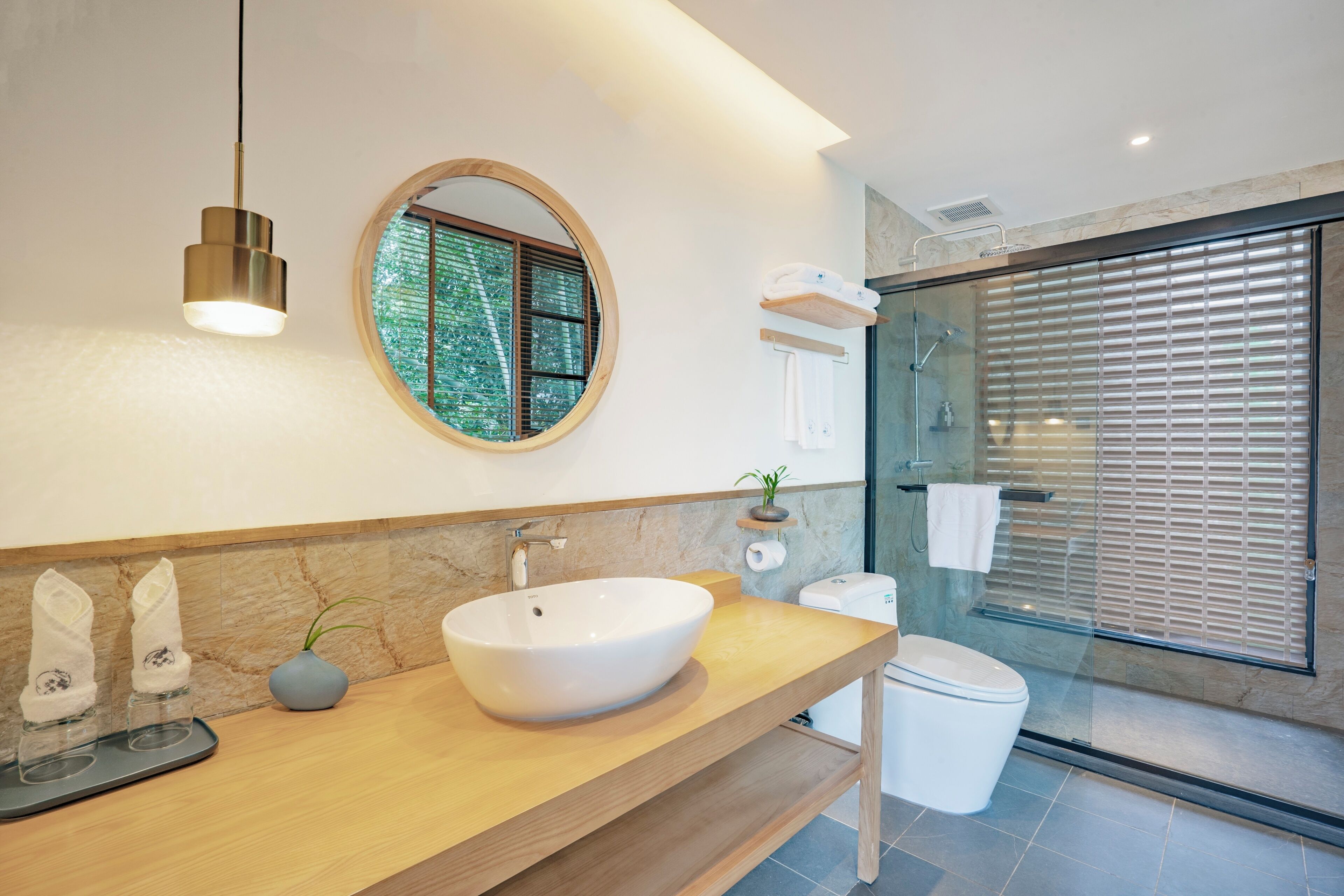 A modern bathroom with a round mirror, a vessel sink, a toilet, and a glass shower enclosure.