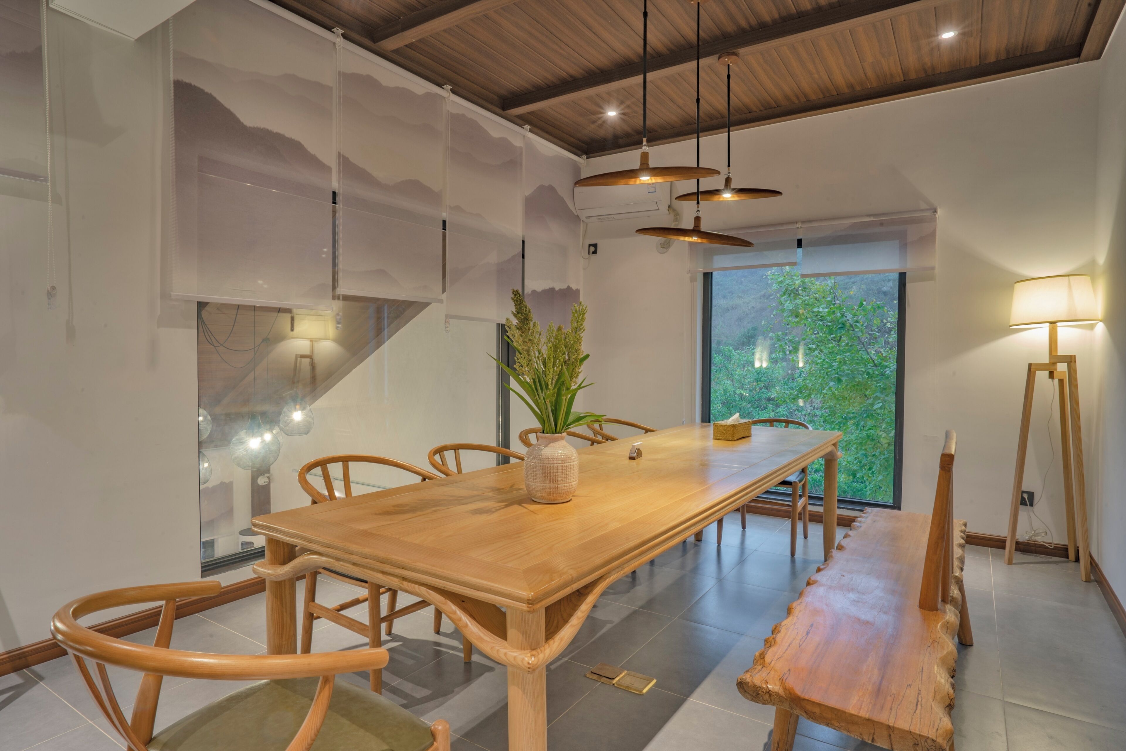 A dining area with a wooden table, chairs, a vase with a plant, pendant lights, and a view through a large window.