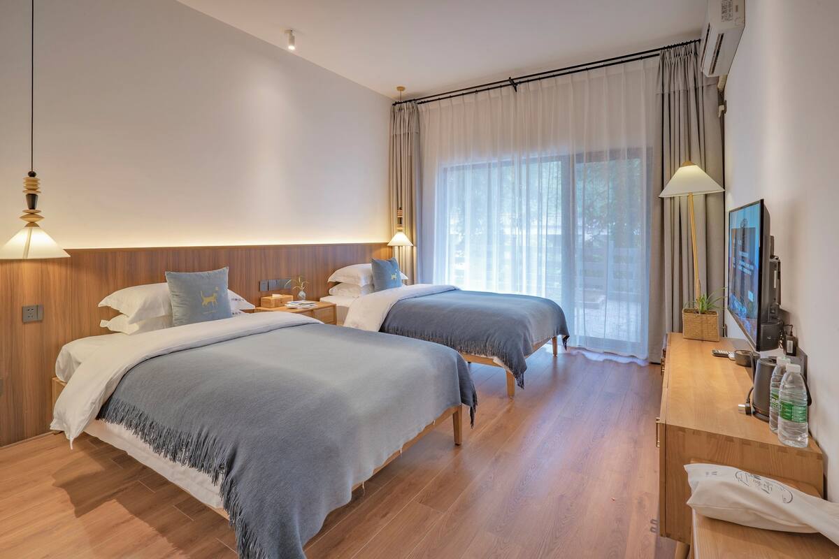 Hotellihuone, jossa on kaksi sänkyä, puinen työpöytä, televisio ja suuri ikkuna, jossa on verhot.