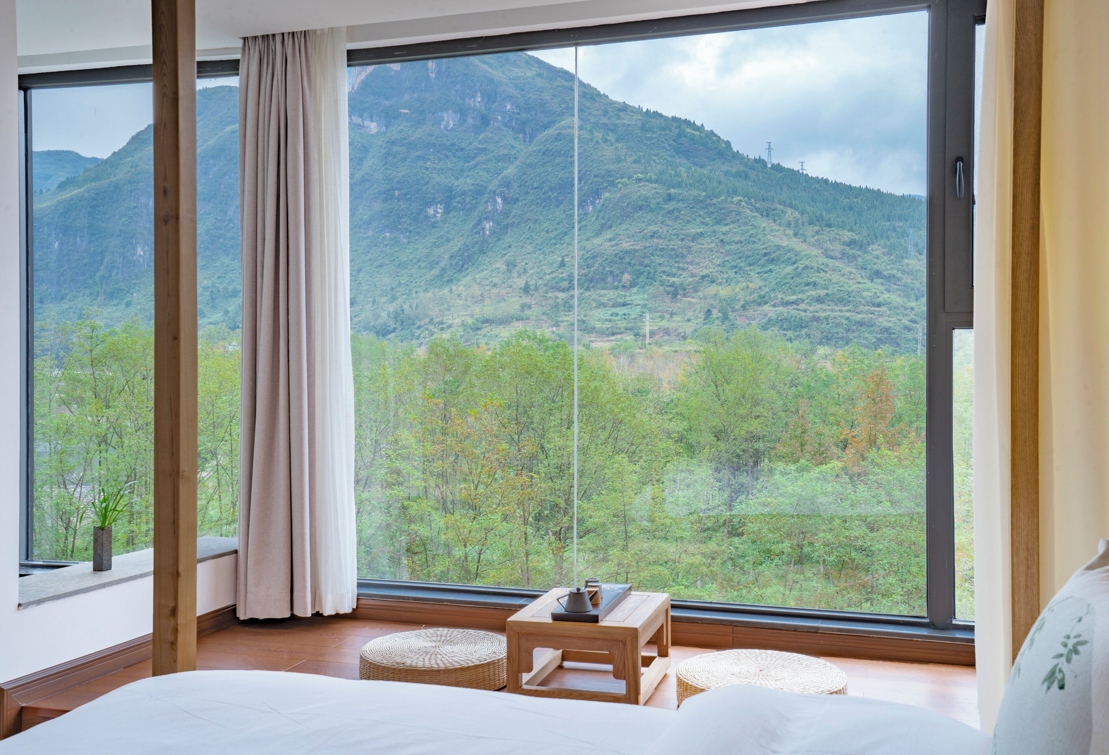 Un dormitorio con un gran ventanal, una cama, una mesita de madera y vistas a un paisaje montañoso.