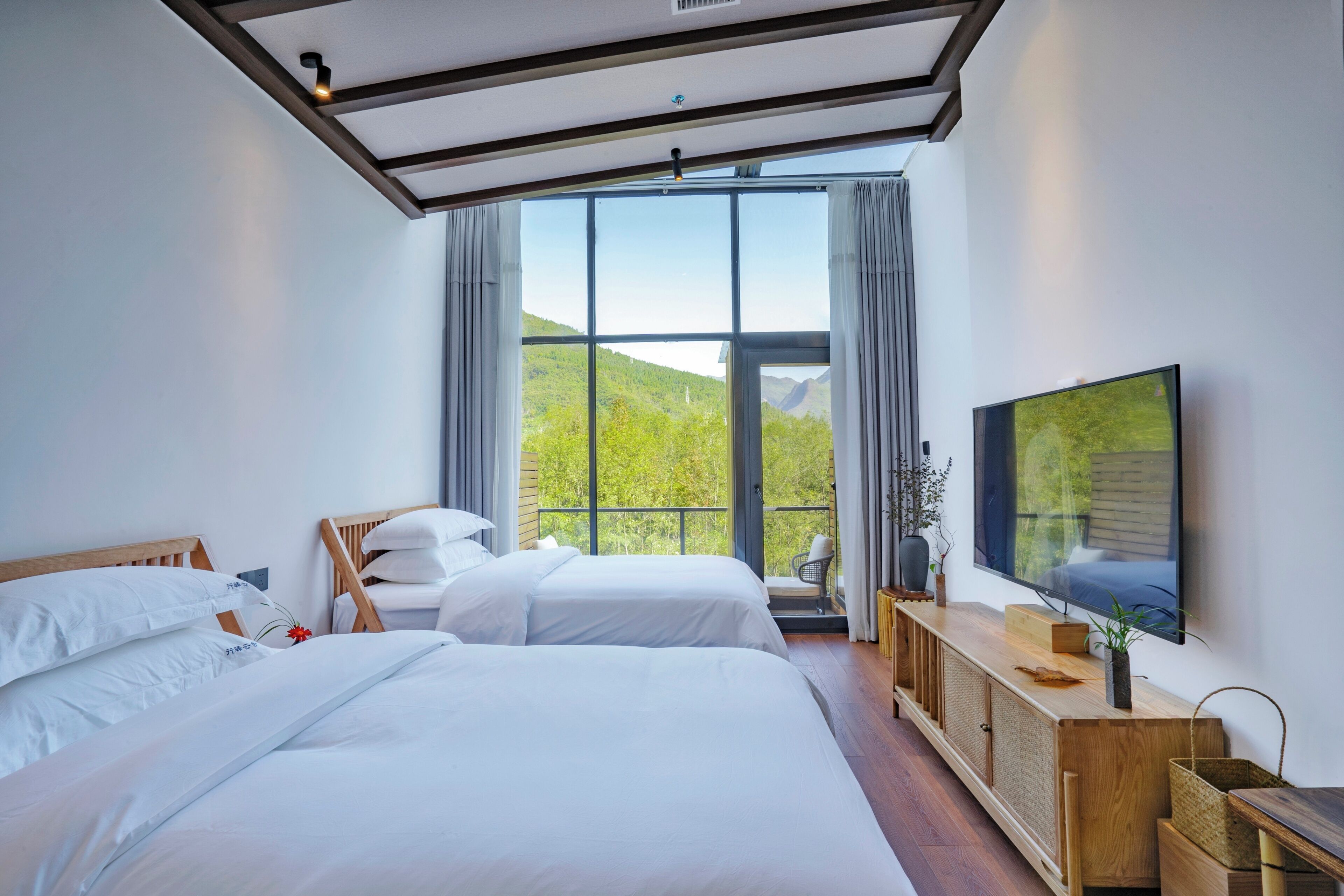 A modern hotel room with two beds, a large window with a view of greenery, a flat-screen TV, and a wooden console table.