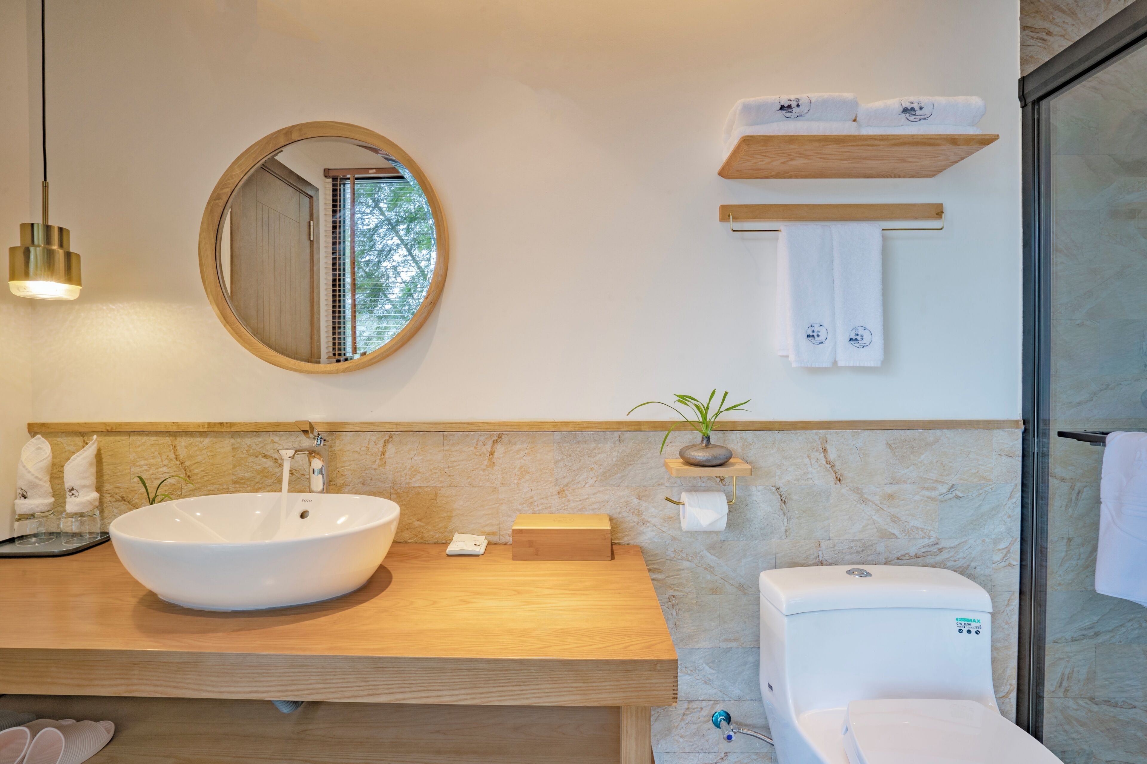 Un baño moderno con un lavamanos ovalado grande, un espejo redondo, un mueble de tocador de madera, un inodoro y una cabina de ducha con mampara de vidrio.