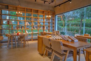 A modern library with wooden bookshelves, tables, and chairs, large windows with a view of greenery.