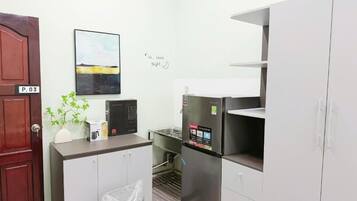 Uma cozinha compacta com geladeira, micro-ondas e uma pia pequena. Há uma porta com a inscrição 'P.03' e uma obra de arte fixada na parede.
