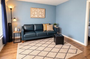 A living room with a blue wall, a black leather sofa, a patterned rug, a floor lamp, and a small table with a vase. - Cozy 1BR 1BA Upstairs Unit with Fast WiFi & Smart HDTV Near Downtown Bay City! (Bay City)