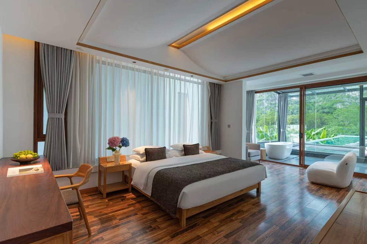 Ein modernes Hotelzimmer mit einem großen Bett, einem Schreibtisch, einem Sessel, einer Couch und Blick auf einen üppigen Garten durch große Fenster.