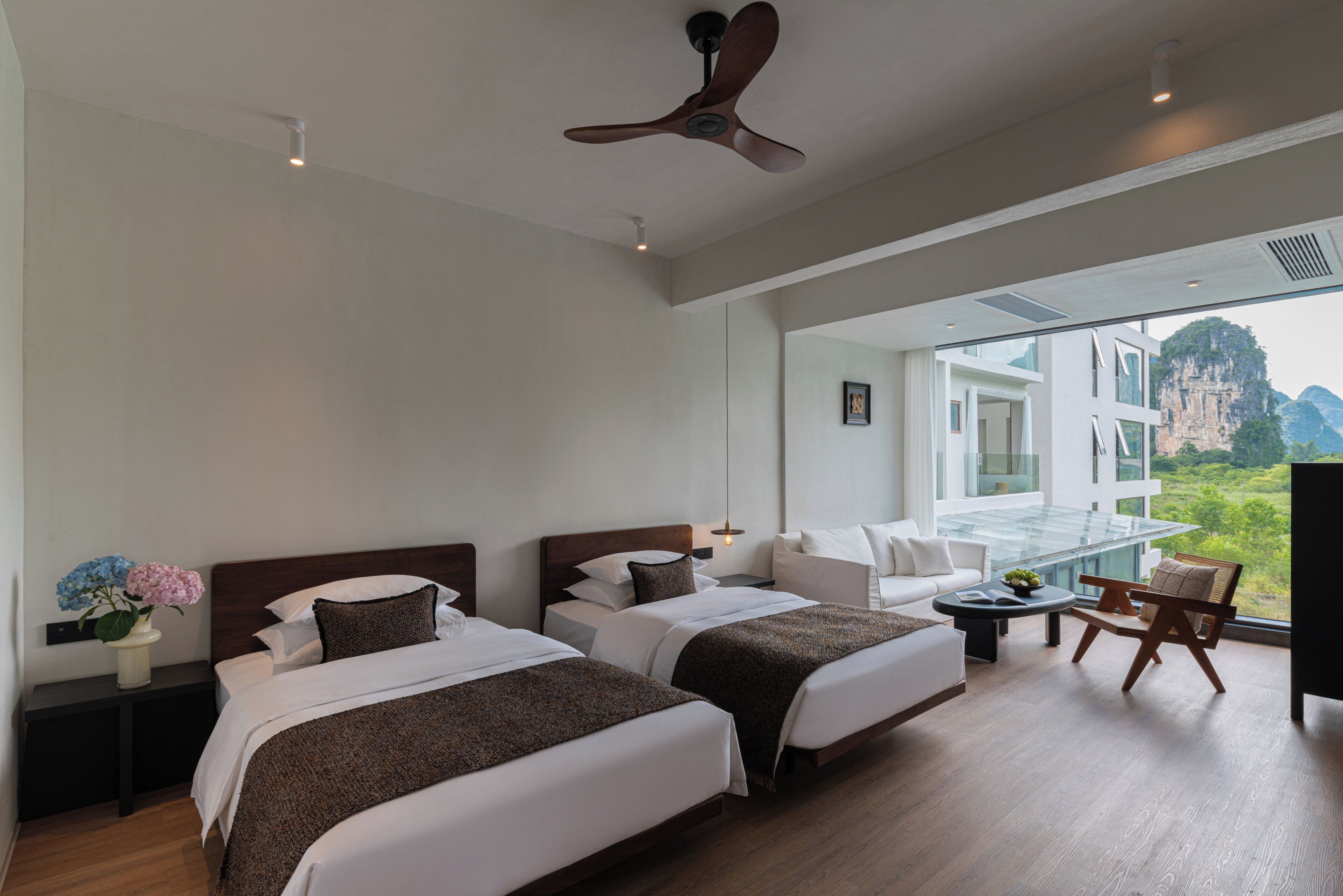 Quarto de hotel com duas camas, um sofá, uma mesa de centro, um ventilador de teto e uma grande janela com vista para as montanhas.
