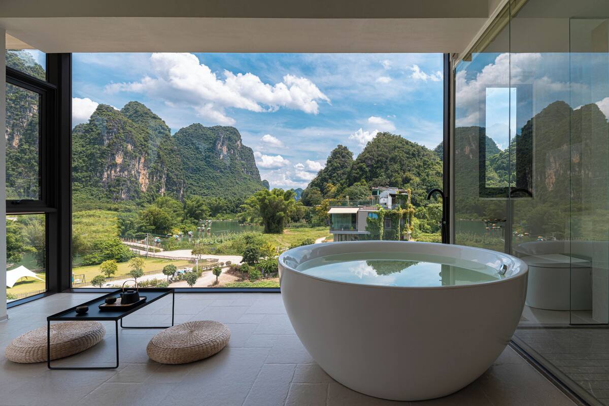 Ein modernes Badezimmer mit einer großen Badewanne, Blick auf eine gebirgige Landschaft und einer kleinen Sitzecke mit Kissen.