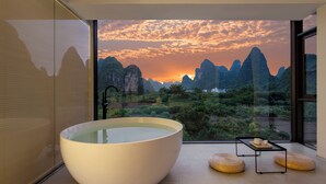 Un baño moderno con una bañera grande, una ducha con mampara de vidrio y vistas a montañas al atardecer.