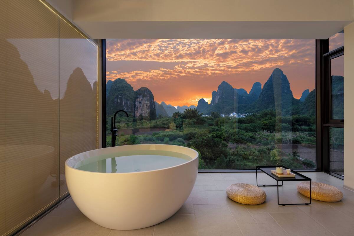 Ein modernes Badezimmer mit einer großen Badewanne, einer Duschkabine aus Glas und Blick auf die Berge bei Sonnenuntergang.