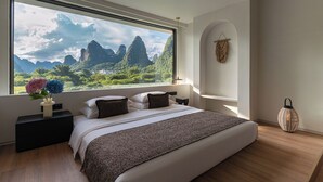 Un dormitorio con un ventanal grande, una cama, una mesita de noche, un macramé colgado en la pared y una lámpara de pie.