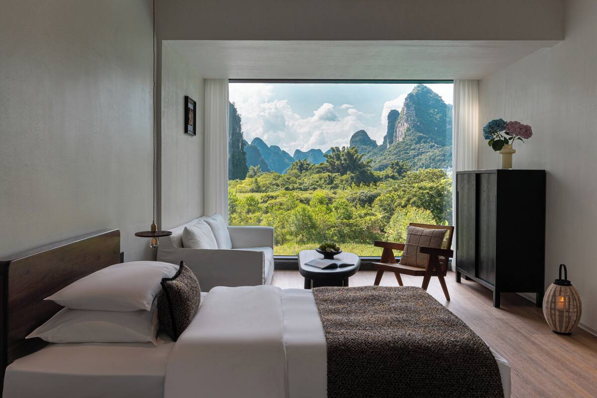 Ein Hotelzimmer mit Bett, Sofa, kleinem Tisch, Stuhl und Blick auf eine gebirgige Landschaft.