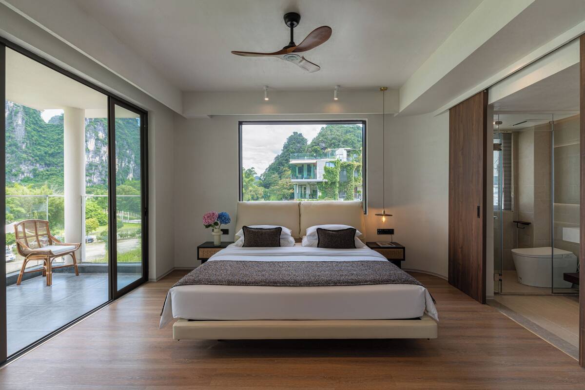 Ein modernes Schlafzimmer mit einem großen Bett, einem Deckenventilator, Blick auf Grünflächen und einer Glasschiebetür, die zu einem Außenbereich führt.