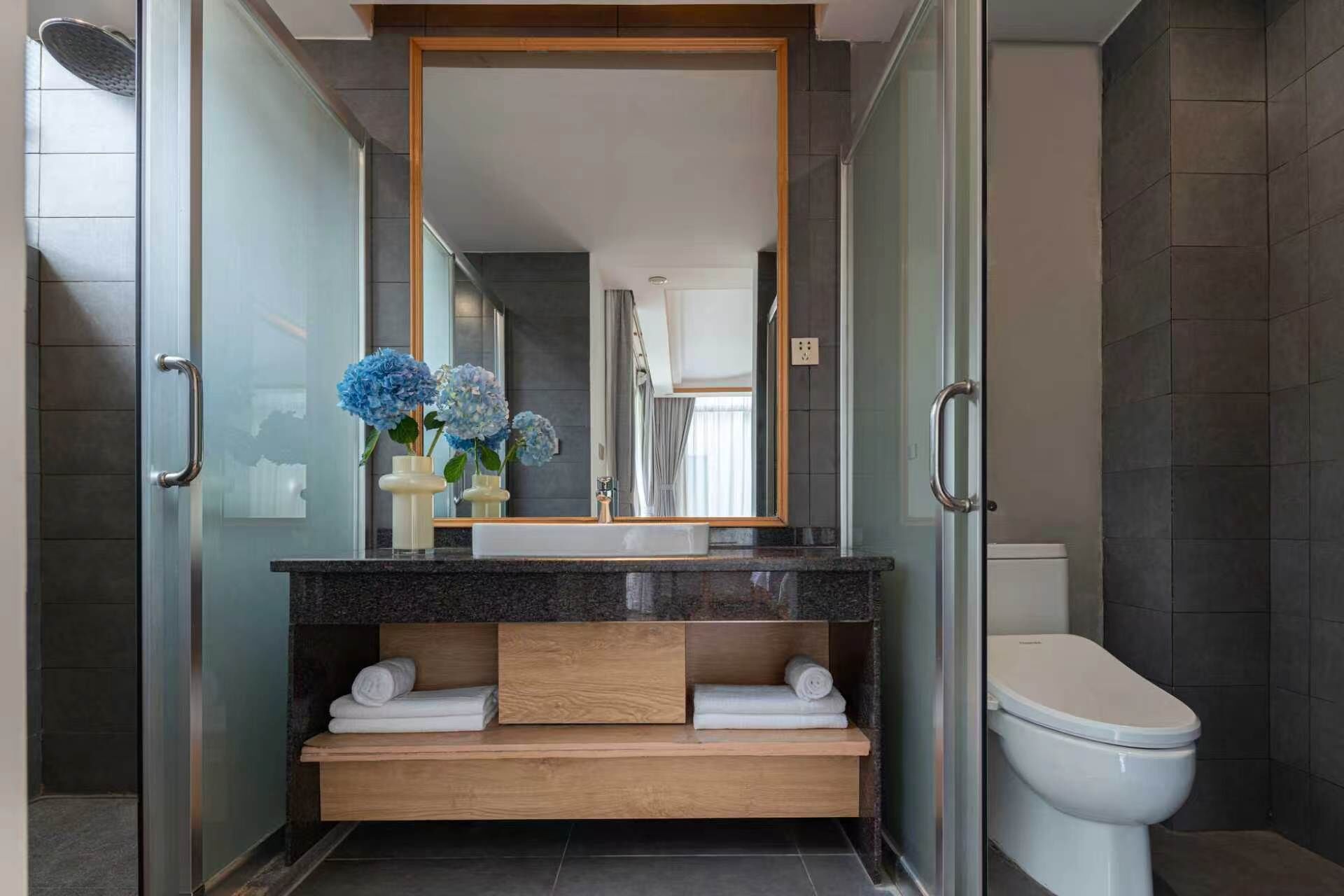 A modern bathroom with a glass shower enclosure, a wooden vanity, a white toilet, and a large mirror.